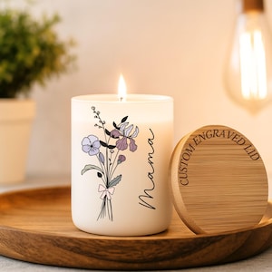 May include: A lit white candle with a floral design and the word "Mama" in script. The candle sits on a wooden tray with a matching lid that reads "CUSTOM ENGRAVED LID". The candle features purple and lavender flowers.