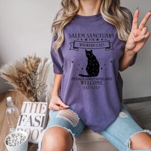 Może przedstawiać: Fioletowy T-shirt z czarnym kotem i tekstem "Salem Sanctuary for Wayward Cats" i "Feral and Familiars Welcome Est. 1692".