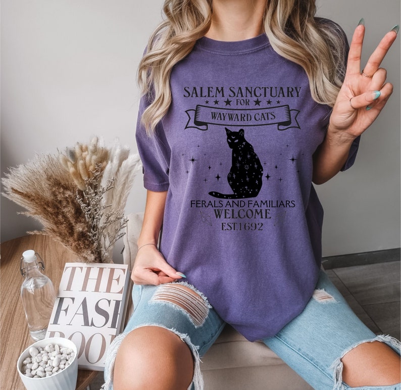 Może przedstawiać: Fioletowy T-shirt z czarnym kotem i tekstem "Salem Sanctuary for Wayward Cats" i "Feral and Familiars Welcome Est. 1692".