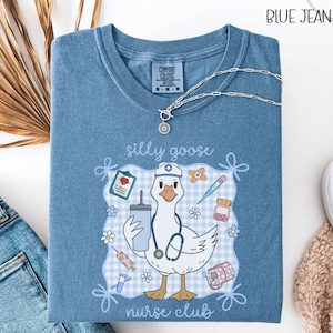 May include: Blue jean colored t-shirt with a graphic of a goose wearing a nurse's hat and stethoscope, holding a drink. The shirt has the words "silly goose nurse club" and medical-themed illustrations.