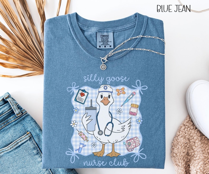 May include: Blue jean colored t-shirt with a graphic of a goose wearing a nurse's hat and stethoscope, holding a drink. The shirt has the words "silly goose nurse club" and medical-themed illustrations.