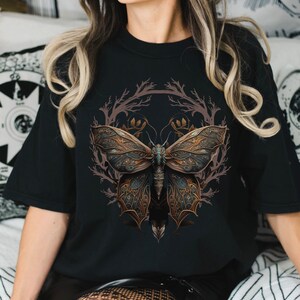 May include: A black t-shirt with a graphic of a detailed, intricate butterfly with gold and blue accents. The butterfly is surrounded by a circular design of brown branches.