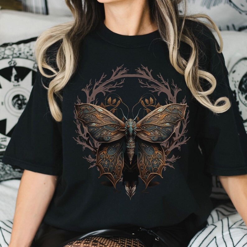 May include: A black t-shirt with a graphic of a detailed, intricate butterfly with gold and blue accents. The butterfly is surrounded by a circular design of brown branches.