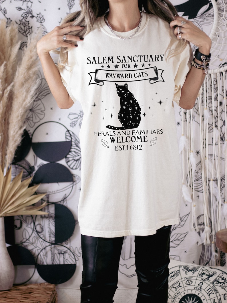Może przedstawiać: Biały t-shirt z grafiką czarnego kota i tekstem "Salem Sanctuary for Wayward Cats. Feral and Familiars Welcome. Est. 1692."