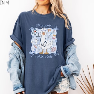 May include: Denim blue t-shirt with a graphic of a cartoon goose wearing a nurse's hat and stethoscope, holding a drink and surrounded by medical supplies. The text "silly goose nurse club" is displayed on the shirt.