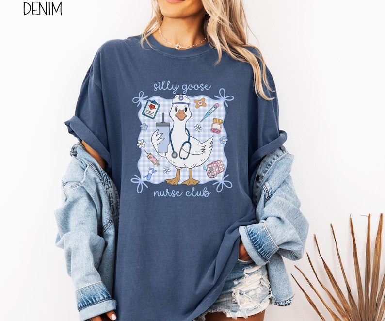 May include: Denim blue t-shirt with a graphic of a cartoon goose wearing a nurse's hat and stethoscope, holding a drink and surrounded by medical supplies. The text "silly goose nurse club" is displayed on the shirt.