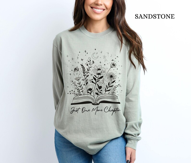 May include: Sandstone-coloured long-sleeve shirt with a black floral and book graphic. The graphic features an open book with flowers and the text "Just One More Chapter". The shirt is a neutral colour.