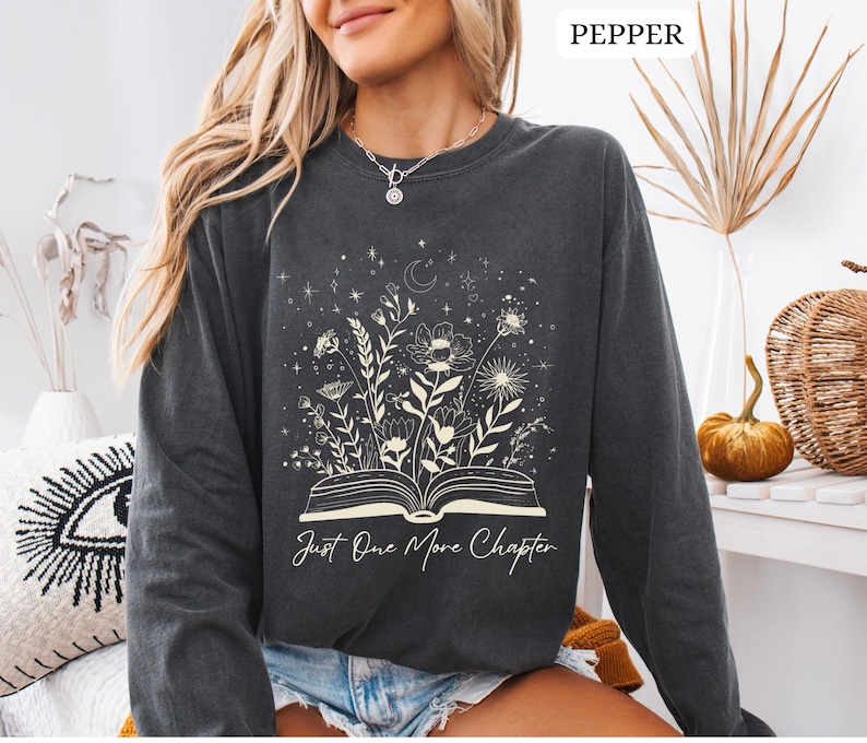 May include: A dark grey long-sleeved sweatshirt with a floral and celestial design. The graphic features an open book with flowers and stars, and the text "Just One More Chapter". The sweatshirt is a comfortable, casual style.