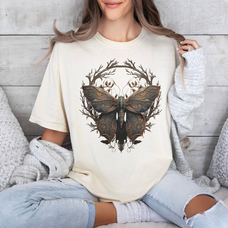 May include: A cream-colored t-shirt with a graphic of a detailed, intricate butterfly with brown, gold, and blue accents. The butterfly is surrounded by a wreath of stylized branches.