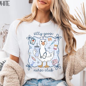 May include: White t-shirt with a cartoon goose wearing a nurse's hat and stethoscope, surrounded by medical-themed illustrations. The text "silly goose nurse club" is displayed above and below the goose. The word "WHITE" is in the upper left corner.