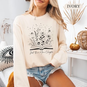 May include: Cream-coloured sweatshirt with a floral and celestial design emerging from an open book. The text "Just One More Chapter" is below the illustration. The sweatshirt is long-sleeved and has a crew neck.