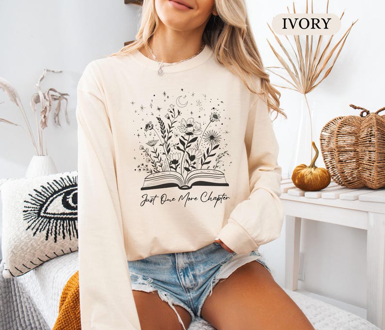 May include: Cream-coloured sweatshirt with a floral and celestial design emerging from an open book. The text "Just One More Chapter" is below the illustration. The sweatshirt is long-sleeved and has a crew neck.