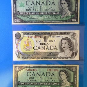 May include: Three Canadian one-dollar banknotes displayed in a protective sleeve. The bills feature portraits of a woman and the text "Canada" and "One Dollar". The top bill commemorates the Centennial of Canadian Confederation.