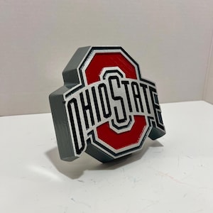 May include: A three-dimensional Ohio State University logo sign featuring a red block O with a white border and the word OHIO STATE in black lettering. The sign has a thick gray outer edge and a layered design with a textured finish.