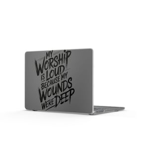 Puede incluir: Una pegatina para portátil de color gris oscuro con tipografía negra estilo pincel que dice: MY WORSHIP IS LOUD BECAUSE MY WOUNDS WERE DEEP. El texto está dispuesto en un diseño apilado y estilizado sobre la tapa de un portátil cerrado.