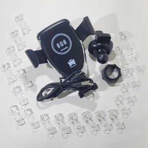 Puede incluir: Un soporte de teléfono para coche negro y plateado con una almohadilla de carga inalámbrica. El soporte tiene una cabeza giratoria de 360 grados y una base de ventosa. La almohadilla de carga tiene un círculo blanco con el texto "Fast Charge" y un icono de coche. El soporte viene con un cable USB y un juego de clips de plástico transparentes.