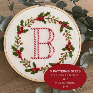 May include: Embroidered letter "B" in red and white, surrounded by a wreath of green leaves and red berries, set in a wooden embroidery hoop. The image includes text: "5 PATTERNS SIZES Includes all letters A-Z Plus numbers 0-9."