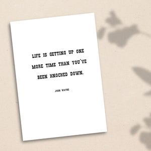 May include: A white printable quote card with black text that reads "Life is getting up one more time than you've been knocked down."  The quote is attributed to John Wayne.