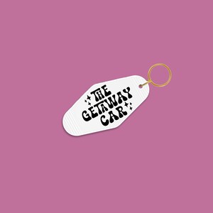 White keychain against a dark pink background, featuring the phrase The Getaway Car surrounded by black vinyl star designs.