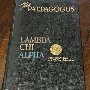 May include: A vintage book titled "The Paedagogus" with "Lambda Chi Alpha" in gold and blue lettering. The cover is dark blue with a textured pattern and a gold seal. The text "...your college home...a lifetime fellowship" is also visible.