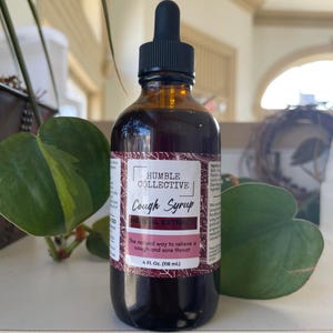Peut inclure: Une bouteille en verre brun de sirop contre la toux Humble Collective avec un bouchon compte-gouttes noir. L'étiquette indique "Cough Syrup, Herbal Extract" et "The natural way to relieve a cough and sore throat." La bouteille contient 118 ml.