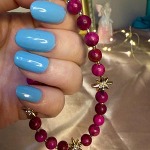 May include: A beaded bracelet featuring alternating deep pink and burgundy round beads. Gold-colored star charms with small clear crystals are interspersed throughout the bracelet. The bracelet is held up against a soft, pinkish background.