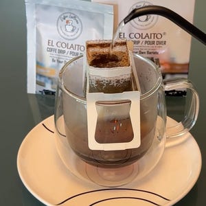 May include: A clear glass mug with a coffee drip filter is set on a white saucer. The filter is being filled with hot water from a black gooseneck kettle. The coffee is labeled "EL COLAITO COFFE DRIP/POUR". A box with the same branding is in the background.
