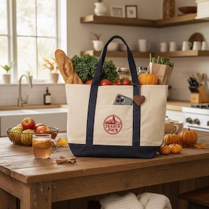 May include: A canvas tote bag with navy blue accents, filled with groceries. The bag features the "Trader Joe's" logo and a pocket holding a phone. A heart-shaped charm hangs from the handle. The bag sits on a wooden table with pumpkins and apples.