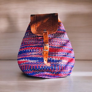 Op de afbeelding: Een kleine, rood, blauw en witte geweven rugzak met een bruin leren klep en een gespsluiting.
