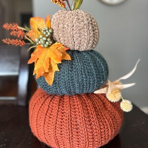 May include: A decorative, stacked pumpkin sculpture. The bottom pumpkin is rust-colored, the middle is teal, and the top is tan. The pumpkins are adorned with fall foliage and berries, creating a seasonal centerpiece.