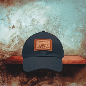 May include: A dark blue baseball cap with a brown leather patch that says "Nature Bound" and features a mountain range and a sun graphic.
