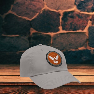 以下が含まれることがあります： 白い鳩と十字架、そして「Love」の文字が描かれた茶色のパッチが付いた灰色のベースボールキャップです。