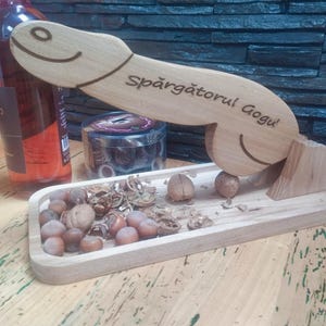 Puede incluir: Un cascanueces de madera con una cara sonriente y el texto "Spargatorul Gogu" grabado. El cascanueces está sobre una bandeja de madera con nueces y avellanas. Una botella de vino es visible en el fondo.
