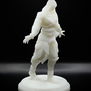 May include: White 3D printed figurine of a person wearing a hooded cloak and holding their hands out. The figurine is standing on a round base.