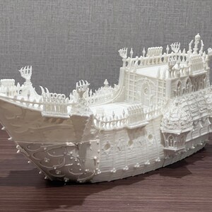 May include: A white, detailed model of a ship with intricate carvings and a gothic design. The ship has a tall, pointed bow and a large, ornate stern.