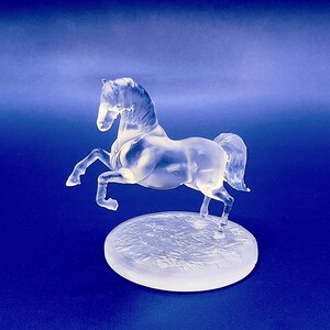 May include: A clear glass horse figurine standing on a textured base. The horse is in a rearing pose with its head turned to the side.