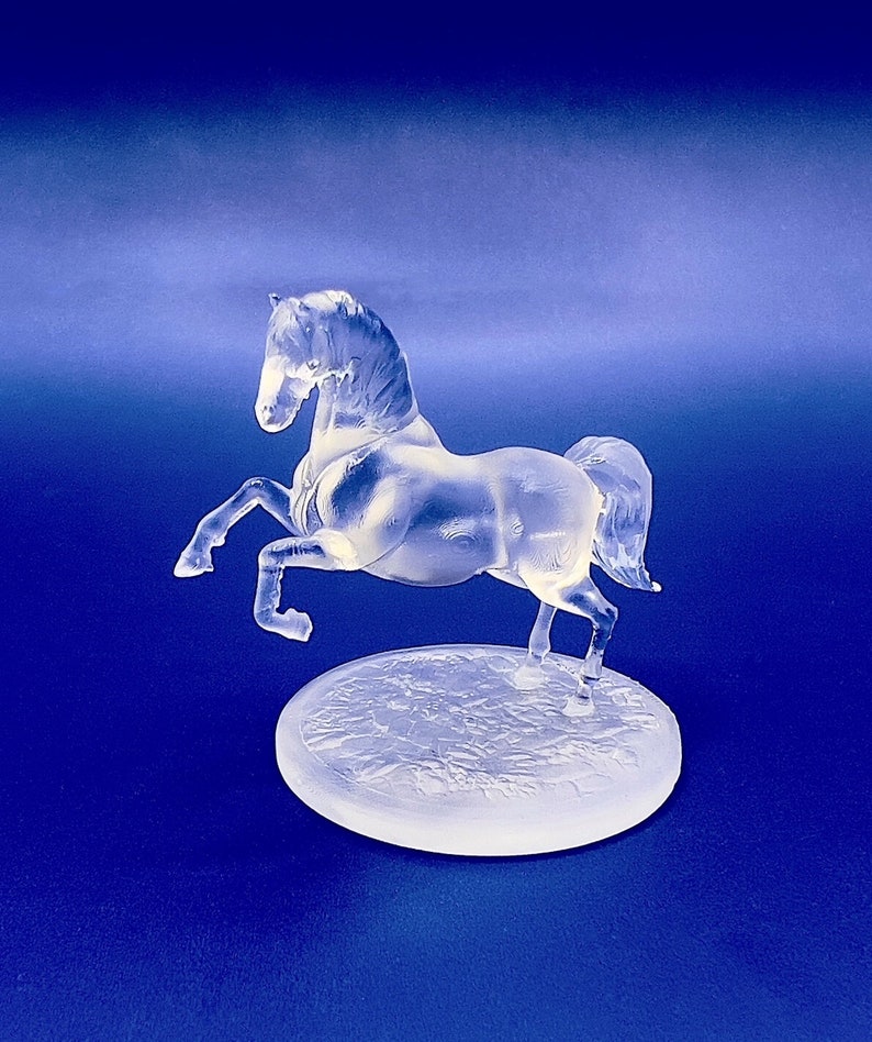 May include: A clear glass horse figurine standing on a textured base. The horse is in a rearing pose with its head turned to the side.