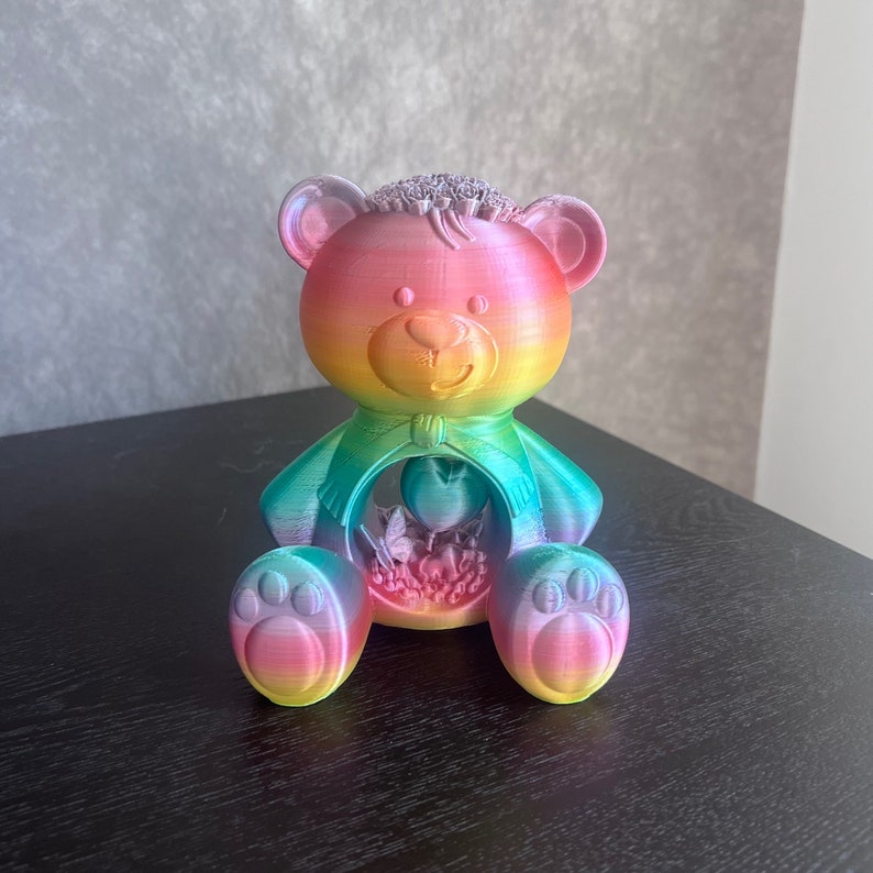 May include: A rainbow-colored teddy bear with a heart and flowers inside its chest. The bear is made of 3D printed plastic and has a glossy finish.