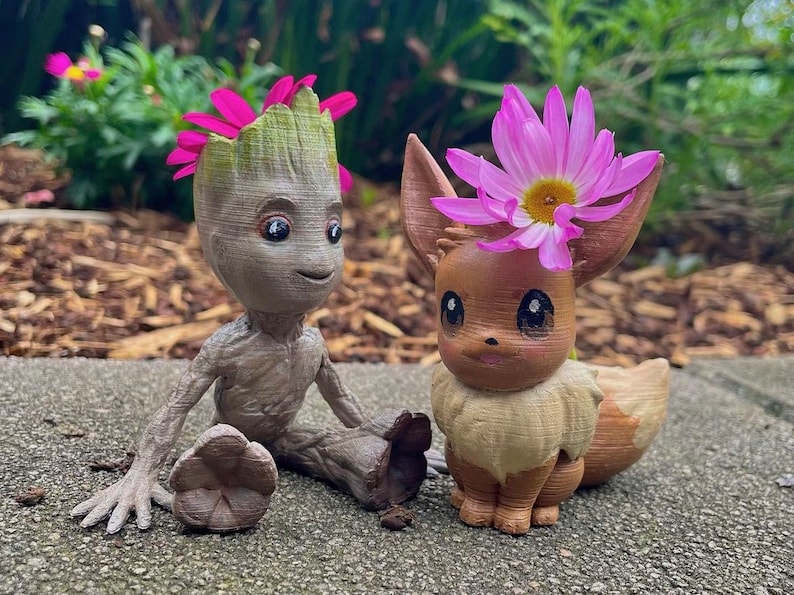 May include: A brown and gray Groot figurine with pink flowers on its head, sitting next to a brown Eevee figurine with a pink flower on its head.
