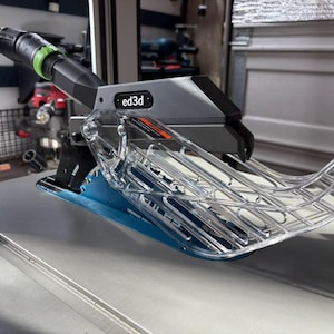 May include: A close-up of a clear plastic dust collection shroud attached to a table saw. The shroud has a gray and black base with the text "ed3d" printed on it.
