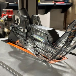 May include: A clear plastic dust collection system for a table saw. The system has a black base with the text "ed3d" printed on it. The system is designed to collect sawdust and other debris from the saw.
