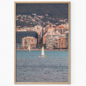 May include: A framed photograph of a cityscape with buildings along the waterfront. Two sailboats are in the water in the foreground. The sky is a light blue color.