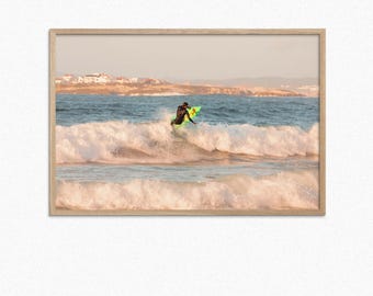 May include: A surfer rides a green and yellow surfboard on a wave with white foam. The surfer is wearing a black wetsuit and is facing the right side of the image. The background is a blue ocean with a sandy beach and a distant coastline.