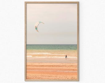 Może przedstawiać: Zdjęcie w ramie przedstawiające osobę uprawiającą kitesurfing na plaży. Latawiec jest w jasnych kolorach niebieskim i białym i lata wysoko w powietrzu. Plaża jest piaszczysta, a woda ma jasnoniebieski kolor.