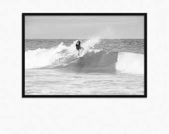 Può includere: Una fotografia in bianco e nero di un surfista che cavalca un'onda. Il surfista indossa una muta e sta cavalcando una tavola da surf. L'onda sta rompendo e l'acqua sta schizzando.