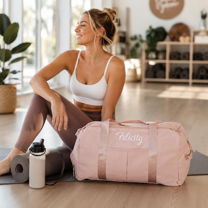 May include: A light pink duffel bag with the name Felicity embroidered in white script on the front pocket. The bag features two sturdy top handles, a zippered main compartment, and a front slip pocket, all constructed from a smooth fabric.