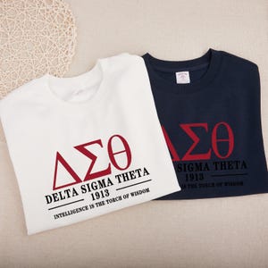 May include: Two sweatshirts, one white and one navy blue, featuring the Greek letters ΔΣΘ in red. The text "DELTA SIGMA THETA 1913 INTELLIGENCE IS THE TORCH OF WISDOM" is printed below the letters. The other sweatshirt has the same design.