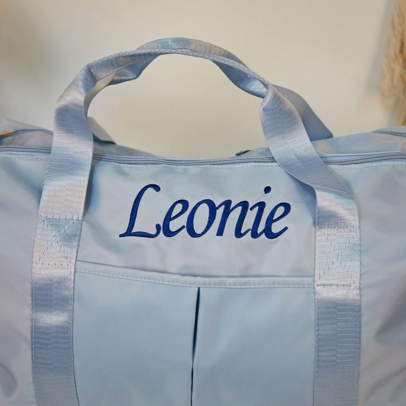 May include: A light blue duffel bag featuring the name Leonie embroidered in dark blue script lettering. The bag includes matching light blue fabric handles and a zippered top closure, with a smooth, solid-colored exterior finish throughout.