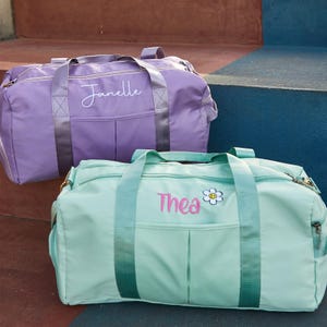 May include: Two personalized duffel bags. One is lavender with "Janelle" embroidered in white script. The other is mint green with "Thea" and a daisy embroidered in pink. Both bags have handles and zippers.