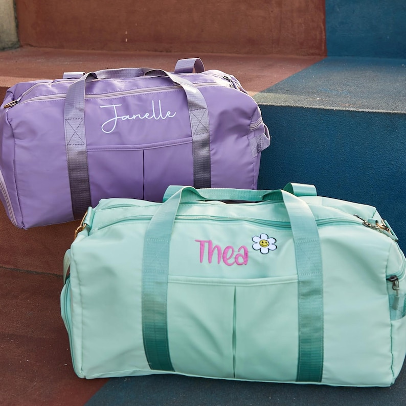 May include: Two personalized duffel bags. One is lavender with "Janelle" embroidered in white script. The other is mint green with "Thea" and a daisy embroidered in pink. Both bags have handles and zippers.